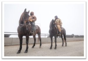 At a feedyard, two cowboys on horses in very cold weather.